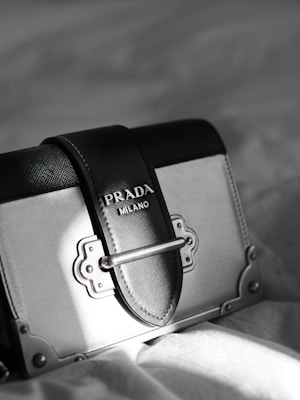 A luxury designer handbag is prominently displayed, featuring a stylish rectangular shape with a sophisticated metal clasp. The bag has a black and gray color scheme with fine stitching details. A Prada Milano logo is elegantly embossed on the leather.