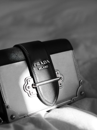 A luxury designer handbag is prominently displayed, featuring a stylish rectangular shape with a sophisticated metal clasp. The bag has a black and gray color scheme with fine stitching details. A Prada Milano logo is elegantly embossed on the leather.
