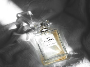A glass perfume bottle with an elegant, rectangular shape lying on a crumpled, light-colored fabric. The label on the bottle reads 'N°5 CHANEL PARIS EAU DE PARFUM'. Light from an unseen source creates bright reflections on the bottle and the fabric, highlighting the transparency and clarity of the glass.