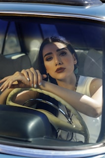 A woman with long dark hair and bold makeup is seated inside a car, resting her arms on the steering wheel and looking thoughtfully out of the front windshield. Sunlight and shadows play across her face and the car's interior. She is wearing a sleeveless white top.
