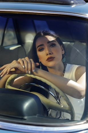 A woman with long dark hair and bold makeup is seated inside a car, resting her arms on the steering wheel and looking thoughtfully out of the front windshield. Sunlight and shadows play across her face and the car's interior. She is wearing a sleeveless white top.