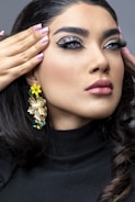 A close-up of a person with meticulously styled hair and makeup, showcasing intricate eye makeup with thick eyeliner and long lashes. The person has their hands touching their temples, revealing light pink nail polish. They are wearing statement jewelry, including large, decorative earrings featuring flowers and gemstones. The clothing is a black turtleneck.
