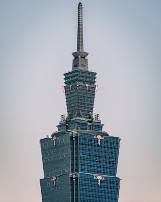 A modern skyscraper with a unique tiered design and a tall spire extends upwards against a clear sky. The building has a sleek, glass facade that reflects sunlight, with intricate details near its upper levels.