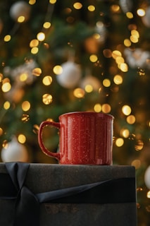 A red mug is placed on top of a gift wrapped with dark paper and a black ribbon. The background is festively decorated with blurred warm yellow lights and white ornaments, suggesting a holiday theme.