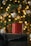 A red mug is placed on top of a gift wrapped with dark paper and a black ribbon. The background is festively decorated with blurred warm yellow lights and white ornaments, suggesting a holiday theme.