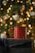 A red mug is placed on top of a gift wrapped with dark paper and a black ribbon. The background is festively decorated with blurred warm yellow lights and white ornaments, suggesting a holiday theme.