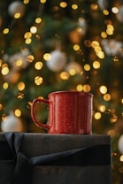 A red mug is placed on top of a gift wrapped with dark paper and a black ribbon. The background is festively decorated with blurred warm yellow lights and white ornaments, suggesting a holiday theme.