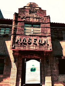 Facade of an old stone building with carved text reading 'MUSEUM' and 'Destileria Limtuaco' above a central window. Below is a mounted information board with a black-and-white photo and text detailing historical information.