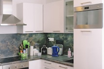 A modern kitchen with white cabinets and a stainless steel oven and range hood. The countertop features various kitchen items including a blue electric kettle, dish soap, a toaster, a paper towel holder, and a small spice rack. The backsplash has a marbled green and beige pattern.