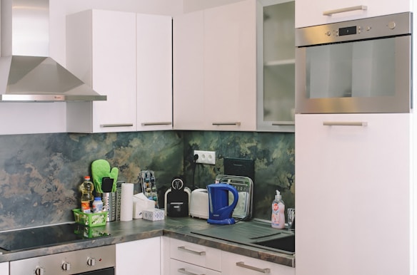 A modern kitchen with white cabinets and a stainless steel oven and range hood. The countertop features various kitchen items including a blue electric kettle, dish soap, a toaster, a paper towel holder, and a small spice rack. The backsplash has a marbled green and beige pattern.