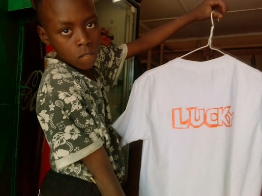 A child is holding a white T-shirt with the word 'LUCKY' printed in bold orange letters. The child is wearing a shirt with a floral pattern and is standing in front of a partially visible green doorway. The interior appears dimly lit with some objects in the background.