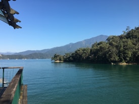 A serene lake surrounded by lush green forests and distant mountains under a clear blue sky. A wooden deck peeks from the left side, giving a sense of calm and tranquility.
