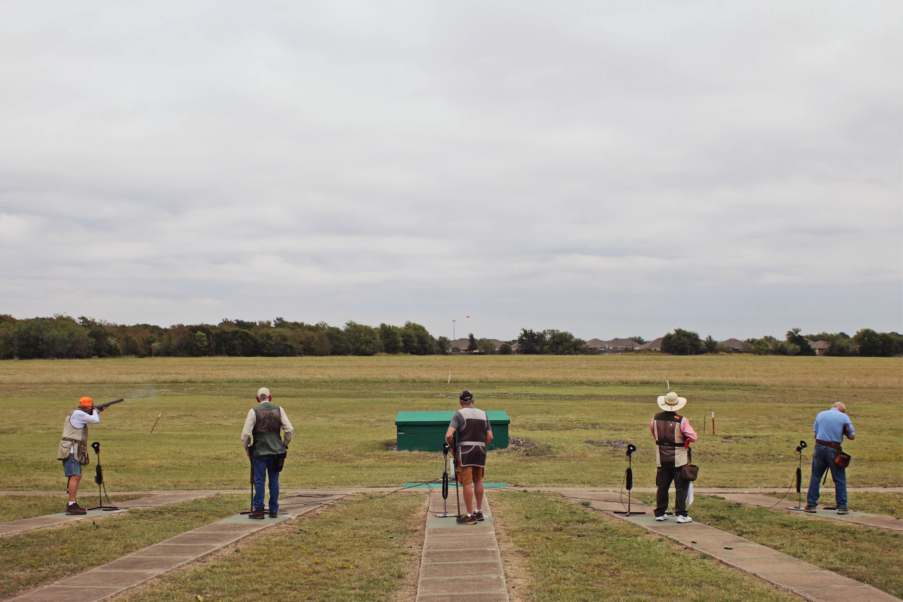 Three people seen from behind on a grass shooting ground under a wide grey sky