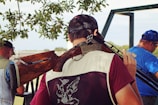 A person wearing a maroon shirt and a mesh vest holds a shotgun over their shoulder. They are standing with their back to the camera, under the shade of a tree, with leaves visible above. In the background, two other individuals are shown, one wearing a blue shirt with ear protection. The setting appears to be outdoors, possibly in a shooting range or hunting area.