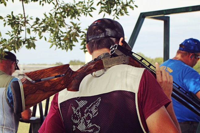 A person wearing a maroon shirt and a mesh vest holds a shotgun over their shoulder. They are standing with their back to the camera, under the shade of a tree, with leaves visible above. In the background, two other individuals are shown, one wearing a blue shirt with ear protection. The setting appears to be outdoors, possibly in a shooting range or hunting area.