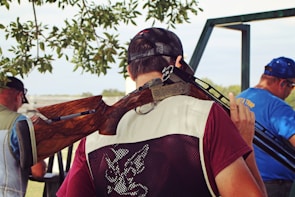 A person wearing a maroon shirt and a mesh vest holds a shotgun over their shoulder. They are standing with their back to the camera, under the shade of a tree, with leaves visible above. In the background, two other individuals are shown, one wearing a blue shirt with ear protection. The setting appears to be outdoors, possibly in a shooting range or hunting area.