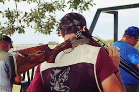A person wearing a maroon shirt and a mesh vest holds a shotgun over their shoulder. They are standing with their back to the camera, under the shade of a tree, with leaves visible above. In the background, two other individuals are shown, one wearing a blue shirt with ear protection. The setting appears to be outdoors, possibly in a shooting range or hunting area.