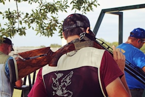 A person wearing a maroon shirt and a mesh vest holds a shotgun over their shoulder. They are standing with their back to the camera, under the shade of a tree, with leaves visible above. In the background, two other individuals are shown, one wearing a blue shirt with ear protection. The setting appears to be outdoors, possibly in a shooting range or hunting area.