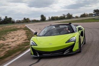 A vibrant green sports car is speeding along a winding track. The vehicle has a sleek, aerodynamic design with a convertible roof and a driver wearing sunglasses and a cap. The background features a partly cloudy sky and some blurred greenery, suggesting motion and speed.