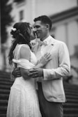 A couple dressed in wedding attire shares an intimate moment. The woman wears a lace dress with floral accents in her hair, while the man is in a light-colored suit with a bow tie. They are embracing on a staircase, suggesting a romantic or wedding setting.