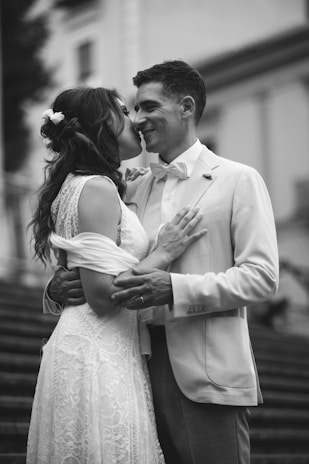 A couple dressed in wedding attire shares an intimate moment. The woman wears a lace dress with floral accents in her hair, while the man is in a light-colored suit with a bow tie. They are embracing on a staircase, suggesting a romantic or wedding setting.