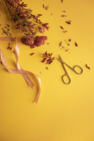 A bouquet of dried pink flowers and leaves is arranged on a bright yellow surface. A pair of silver scissors, gold safety pins, and a pink satin ribbon are also present, adding elements of craft and decoration.