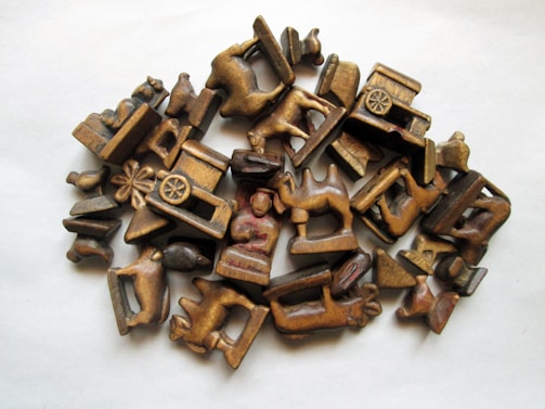 A collection of small, intricately carved wooden figurines and objects, depicting animals, carts, and human figures. The carvings are arranged in a casual pile on a light surface, showcasing a variety of shapes and details in the wood.
