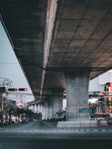 A large concrete overpass stretches across the frame with significant infrastructure visible, including traffic lights and power lines. The scene is urban in nature, featuring trees and sparse vehicular traffic beneath the structure.