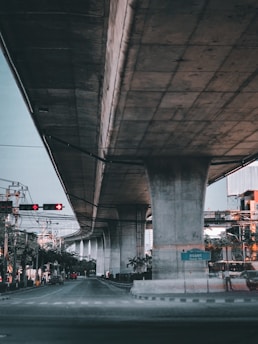 A large concrete overpass stretches across the frame with significant infrastructure visible, including traffic lights and power lines. The scene is urban in nature, featuring trees and sparse vehicular traffic beneath the structure.