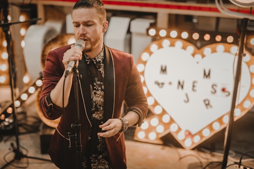 A man sings into a microphone, dressed in a floral-patterned shirt and a burgundy jacket. Behind him, a large illuminated heart-shaped sign with letters is visible. The setting appears to be indoors with warm, soft lighting.