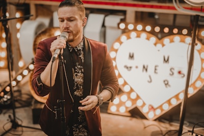 A man sings into a microphone, dressed in a floral-patterned shirt and a burgundy jacket. Behind him, a large illuminated heart-shaped sign with letters is visible. The setting appears to be indoors with warm, soft lighting.