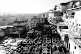 An outdoor tannery scene featuring numerous stone vats used for leather dyeing, surrounded by old, rustic buildings. The monochrome setting highlights textures and structures, with fabrics draped over walls.