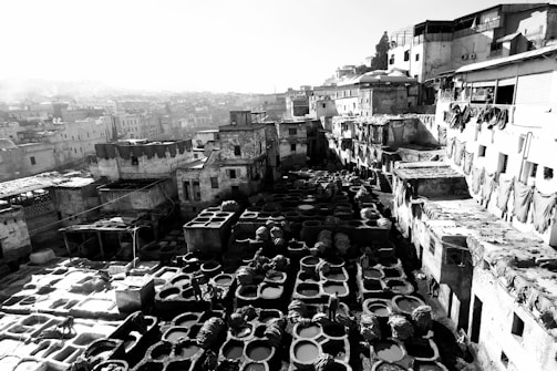 An outdoor tannery scene featuring numerous stone vats used for leather dyeing, surrounded by old, rustic buildings. The monochrome setting highlights textures and structures, with fabrics draped over walls.