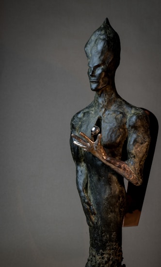 A dark, abstract sculpture with an elongated head and features. Its hand is positioned near its chest, holding a small, spherical object. The surface texture appears rough, and the materials give it a metallic sheen.