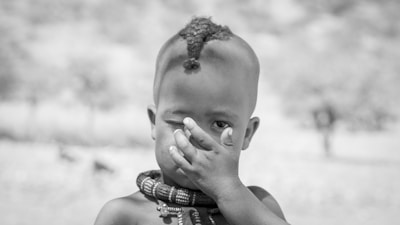 A young child with a distinctive hairstyle stands with one hand covering their face. The child is wearing multiple beaded necklaces, and the background appears to be an outdoor, blurred natural setting.