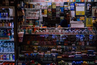 A small convenience store counter is filled with various products like candy, snacks, and cigarettes. Signs are prominently displayed, emphasizing age restrictions on cigarettes and refund policies. To the left, a refrigerated drink display includes bottled beverages and energy drinks. A person is partially visible behind the counter surrounded by lottery tickets and advertisement banners.