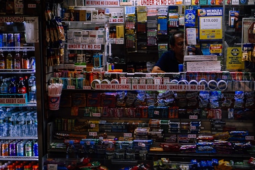 A small convenience store counter is filled with various products like candy, snacks, and cigarettes. Signs are prominently displayed, emphasizing age restrictions on cigarettes and refund policies. To the left, a refrigerated drink display includes bottled beverages and energy drinks. A person is partially visible behind the counter surrounded by lottery tickets and advertisement banners.