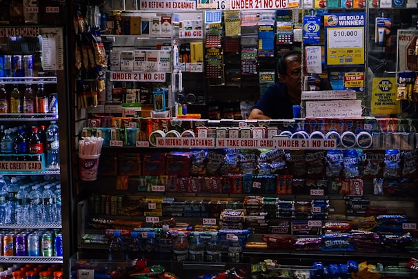 A small convenience store counter is filled with various products like candy, snacks, and cigarettes. Signs are prominently displayed, emphasizing age restrictions on cigarettes and refund policies. To the left, a refrigerated drink display includes bottled beverages and energy drinks. A person is partially visible behind the counter surrounded by lottery tickets and advertisement banners.