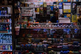 A small convenience store counter is filled with various products like candy, snacks, and cigarettes. Signs are prominently displayed, emphasizing age restrictions on cigarettes and refund policies. To the left, a refrigerated drink display includes bottled beverages and energy drinks. A person is partially visible behind the counter surrounded by lottery tickets and advertisement banners.