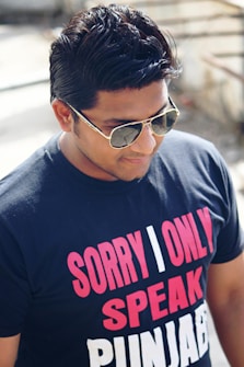 A person wearing sunglasses and a dark blue t-shirt with bold red and white text printed on it, reading 'SORRY I ONLY SPEAK PUNJABI'. The person has neatly styled dark hair and stands in an outdoor setting, with sunlight casting soft shadows.