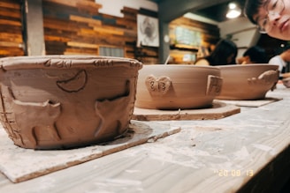 Clay bowls are placed on a wooden table in a pottery studio. The nearest bowl has carvings of animals and symbols, while the others have engraved hearts with numbers. The background features a wooden wall with various artworks and shelves. A person with glasses is partially visible, observing the scene.