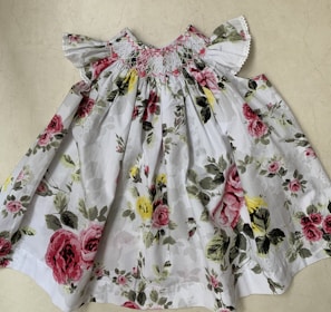 A white floral dress featuring a colorful pattern of pink and red roses with green leaves against a light background. The dress has ruffled short sleeves and detailed smocking near the neckline.