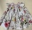 A white floral dress featuring a colorful pattern of pink and red roses with green leaves against a light background. The dress has ruffled short sleeves and detailed smocking near the neckline.