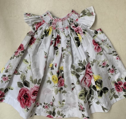 A white floral dress featuring a colorful pattern of pink and red roses with green leaves against a light background. The dress has ruffled short sleeves and detailed smocking near the neckline.