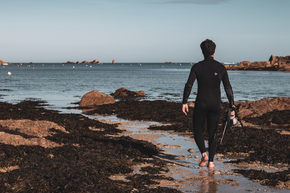 homme en veste noire et pantalon debout sur le rivage rocheux pendant la journée