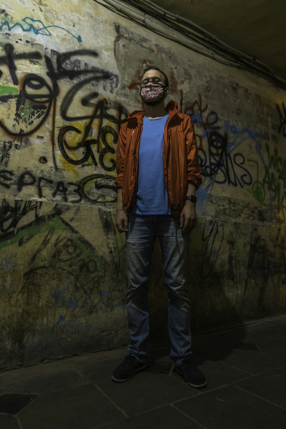 man in orange and gray jacket standing beside wall with graffiti