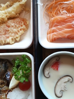 A variety of Japanese dishes are arranged neatly in separate sections. There are slices of salmon sashimi with shredded daikon and carrots. Tempura pieces, including shrimp and vegetables, are lightly battered and fried to a golden color. A bowl of chawanmushi features slices of mushroom and a piece of red fish roe or garnish on top. A grilled fish fillet is paired with a small portion of grated daikon, a cherry tomato, a wedge of lemon, and a sprig of parsley.