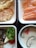 A variety of Japanese dishes are arranged neatly in separate sections. There are slices of salmon sashimi with shredded daikon and carrots. Tempura pieces, including shrimp and vegetables, are lightly battered and fried to a golden color. A bowl of chawanmushi features slices of mushroom and a piece of red fish roe or garnish on top. A grilled fish fillet is paired with a small portion of grated daikon, a cherry tomato, a wedge of lemon, and a sprig of parsley.