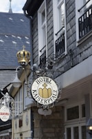 A street view displays signage for 'Hotel Kaiserpfalz' mounted on the exterior of a building with a grey slate roof and multiple windows. The sign has an ornate metal design topped with a decorative element resembling a crown. In the background, there is another sign for 'Sakura Asian Kitchen' also featuring ornate metalwork.