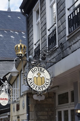 A street view displays signage for 'Hotel Kaiserpfalz' mounted on the exterior of a building with a grey slate roof and multiple windows. The sign has an ornate metal design topped with a decorative element resembling a crown. In the background, there is another sign for 'Sakura Asian Kitchen' also featuring ornate metalwork.